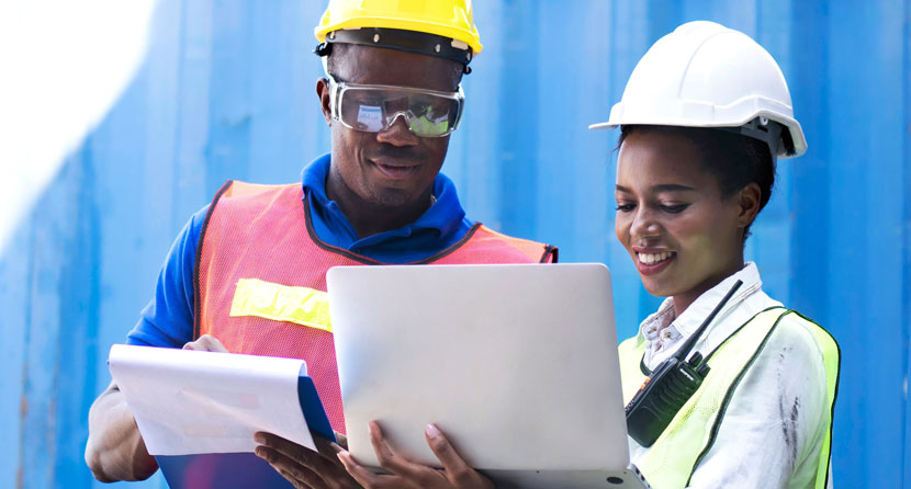 A photograph of workers wearing proper head and eye protection reviewing a plan on a laptop.