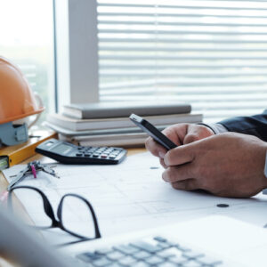 A photograph of a construction project manager looking at a phone at their desk accompanied by a calculator, a laptop, some tools and a helmet.