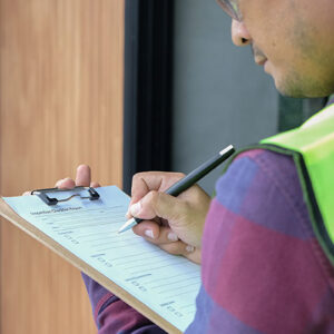 A photograph over the shoulder of a male worker wearing a yellow vest holding a clipboard with a checklist on it.