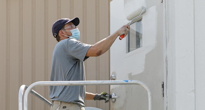Photograph of a man using a paint roller on a mobile office trailer outside of a warehouse.