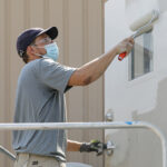Photograph of a man using a paint roller on a mobile office trailer outside of a warehouse.