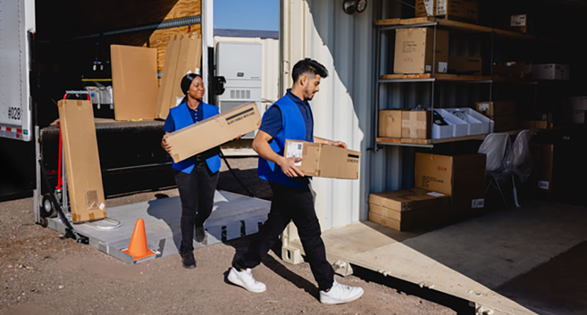 Photograph of two retail employees wearing blue shirts and vests carrying boxes from a truck into a shipping container.