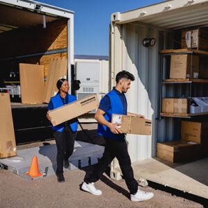 Photograph of two retail employees wearing blue shirts and vests carrying boxes from a truck into a shipping container.